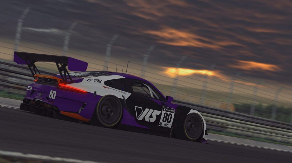 A Porsche 911 GT3 R in an orange, white and purple livery driving in the twilight on a racetrack.