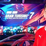A man in a red shirt in front of a background with a big screen saying Gran Turismo 7 on it.