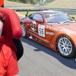 A man with a red shirt and a bright red line in his hair next to a Mercedes racing car in orange and red.